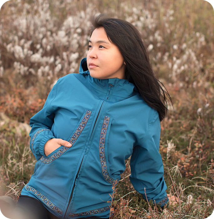 A woman in a teal Naataq Gear jacket with patterned Qaspeq-inspired trim, standing in autumn tundra grass.